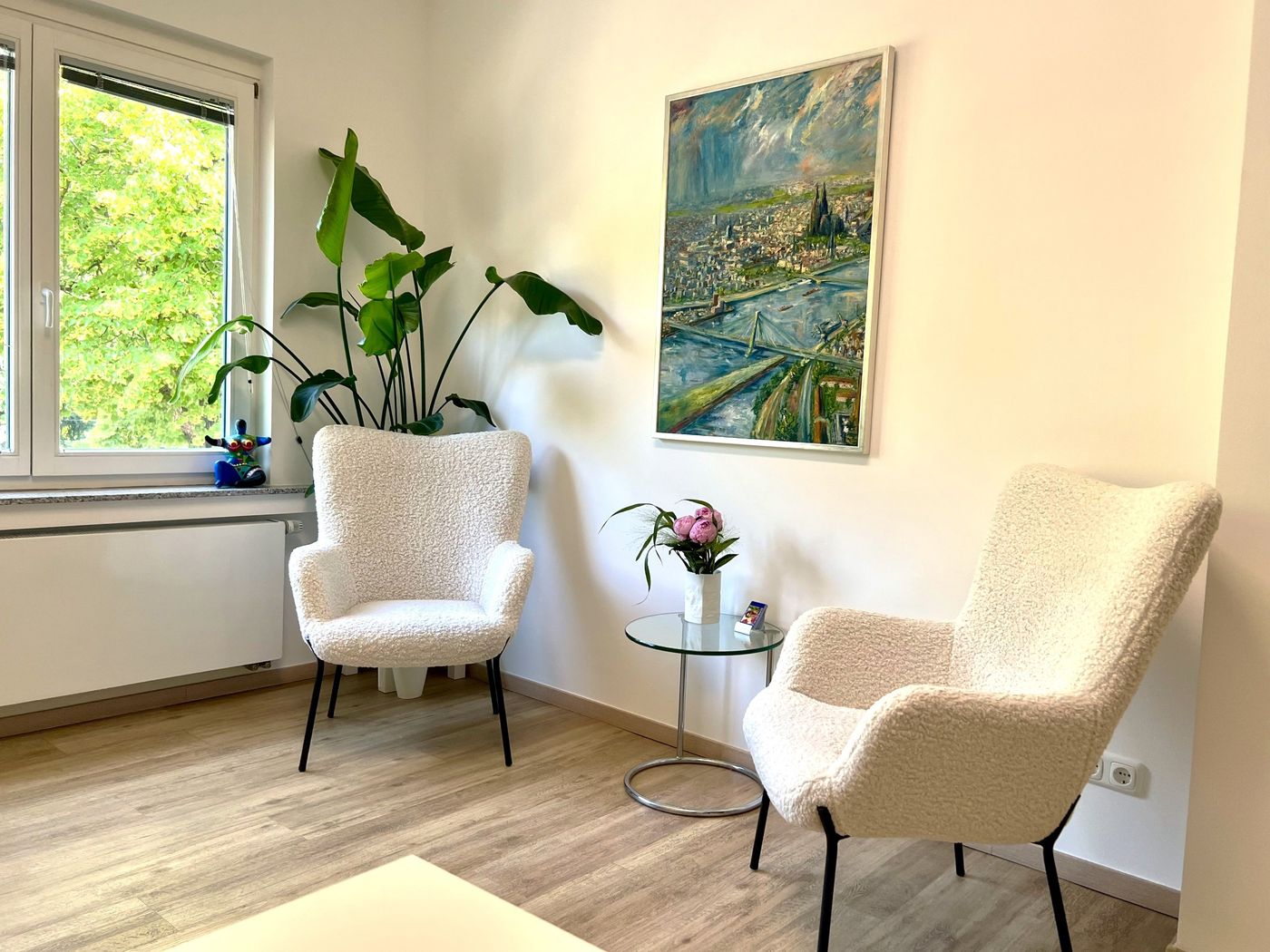 Gesprächsraum der Privatpraxis am Stadtwald, Köln-Lindenthal Heller Gesprächsraum der Privatpraxis mit zwei hellen Sesseln, Grünpflanze und Köln-Gemälde
